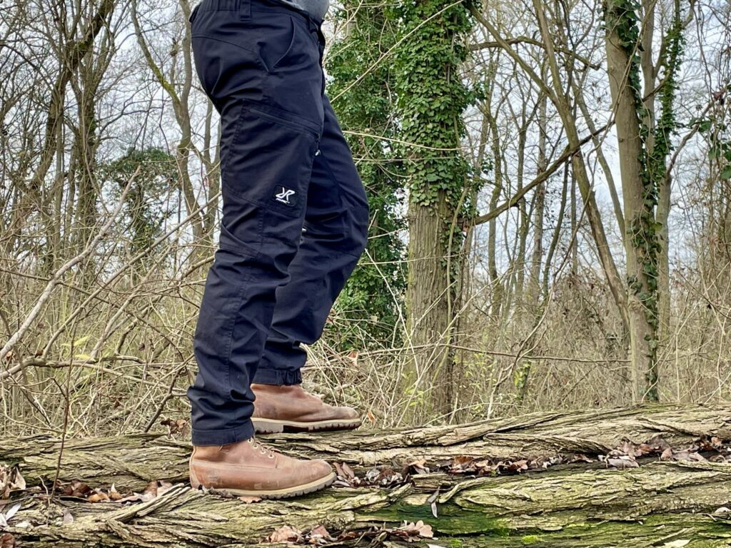 Die Beine einer Person, die die Outdoor Pants von Revolution Race trägt. Wald im Hintergrund.